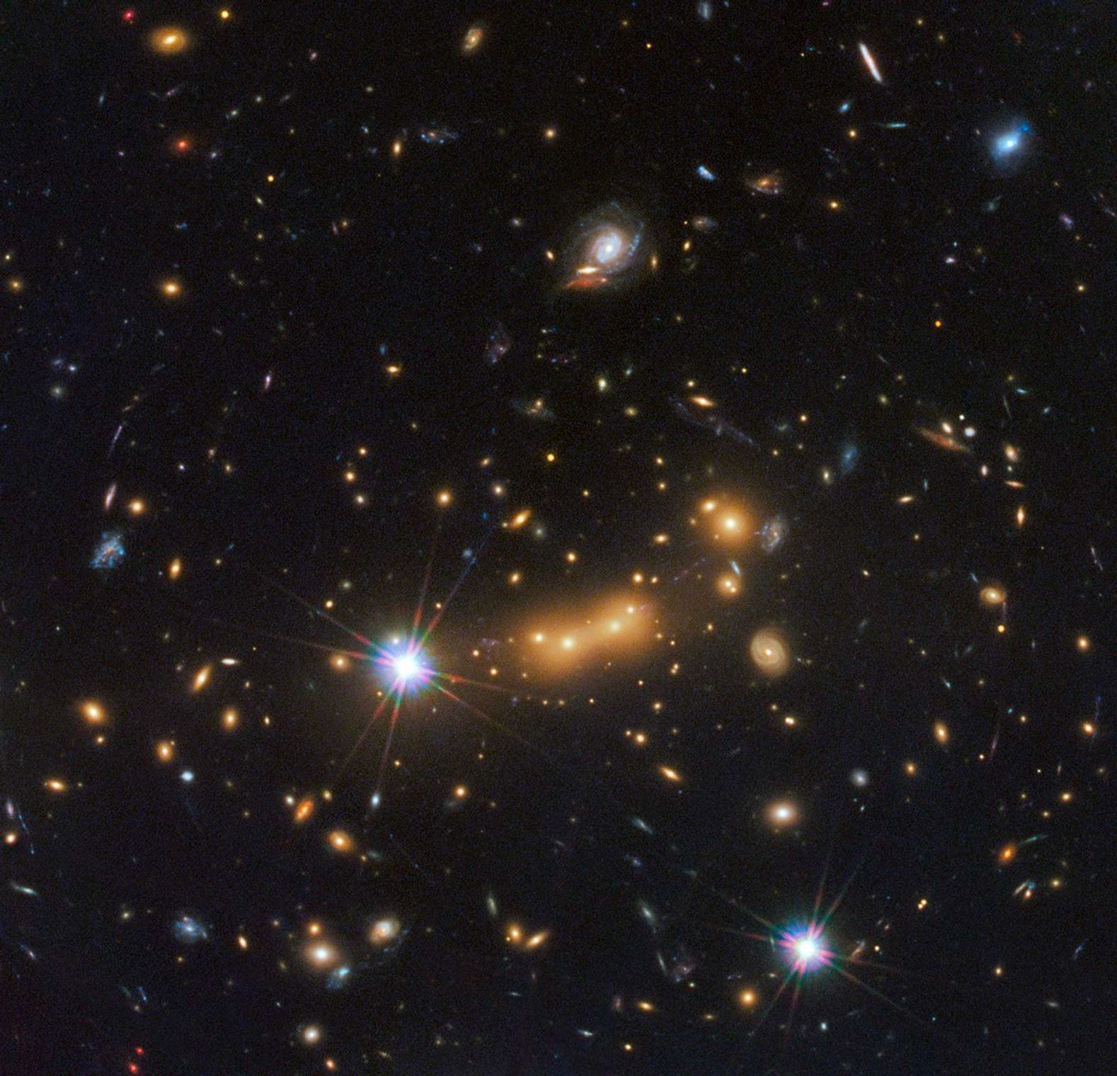 A packed field of galaxies and with faint curved arcs. Large grouping of galaxies warped around what appears to be a giant lens. Each galaxy appears as a glob of light, most of them reddish-orange. Bright blue-white foreground star at left and bottom right.